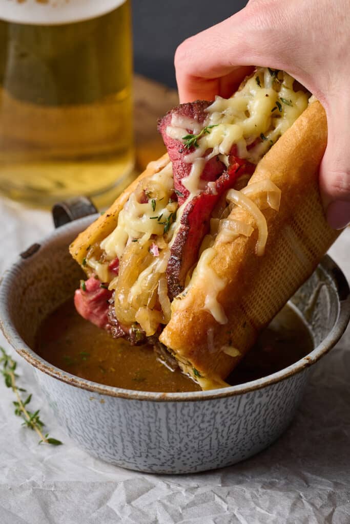 French dip sandwich being dunked into a bowl of au jus sauce.
