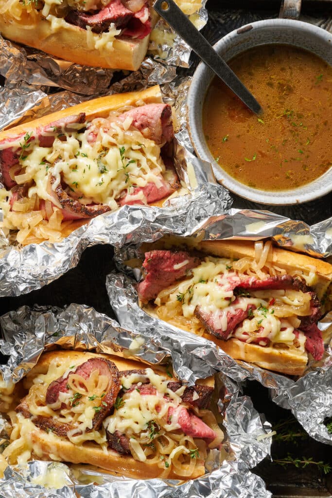 French dip sandwiches wrapped in aluminum foil with au jus on the side.