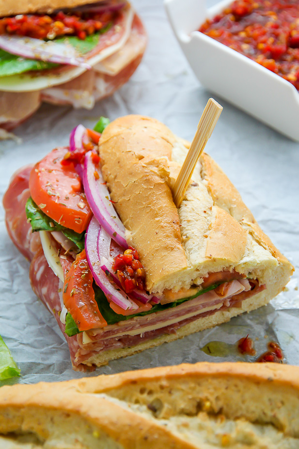 Italian hoagies wit long rolls filled with deli meats, cheese, and veggies topped with olive oil on marble table.