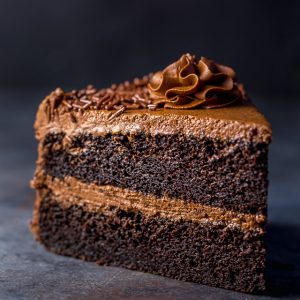 Slice of chocolate cake on a dark table topped with chocolate buttercream frosting.