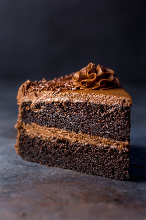 Slice of chocolate cake on a dark table topped with chocolate buttercream frosting.