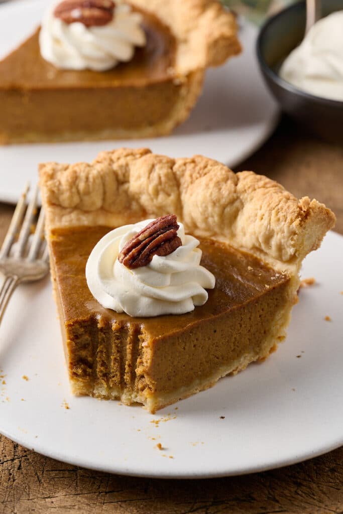 Slice of pumpkin pie on a plate with a bite taken out of it.