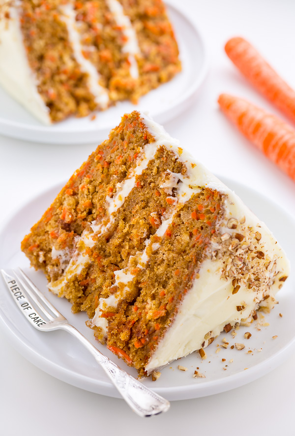 Slice of carrot cake on a dessert plate with a dessert fork and carrots on the side.