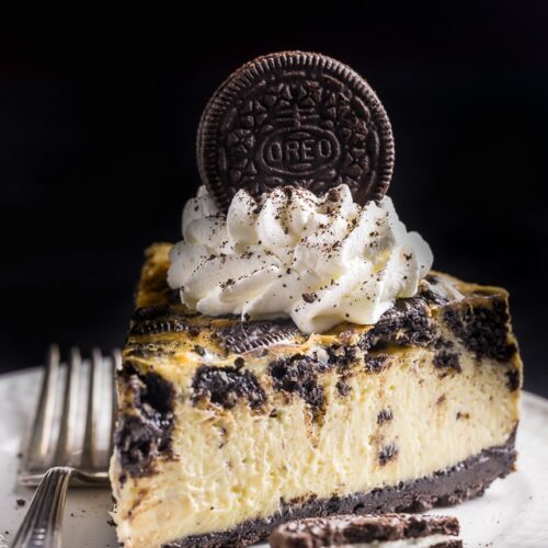 Slice of oreo cheesecake with oreo crust, whipped cream, and an oreo cookie on a white plate.
