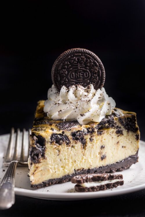 Slice of oreo cheesecake with oreo crust, whipped cream, and an oreo cookie on a white plate.