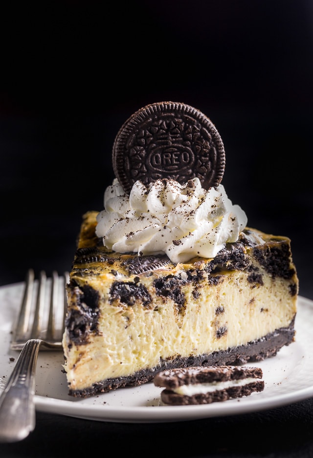 Slice of oreo cheesecake with oreo crust, whipped cream, and an oreo cookie on a white plate.