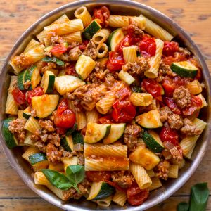 Skillet of rigatoni with tomato sauce, sausage, and zucchini on brown table.