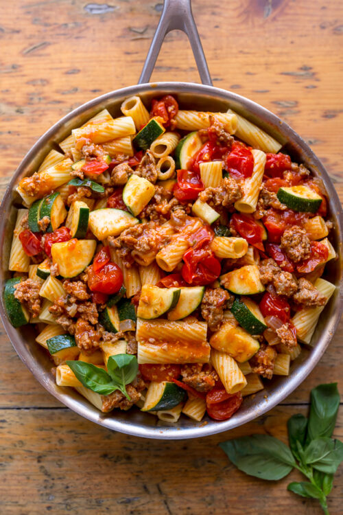 Skillet of rigatoni with tomato sauce, sausage, and zucchini on brown table.