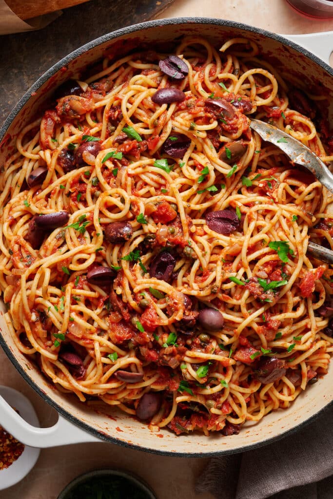 A large pot of pasta puttanesca topped with olives, capers, and parsley.