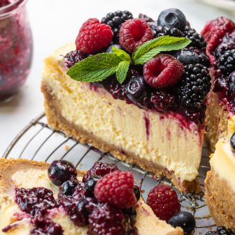 Slice of ricotta cheesecake on a cooling rack topped with fresh berries and berry sauce.