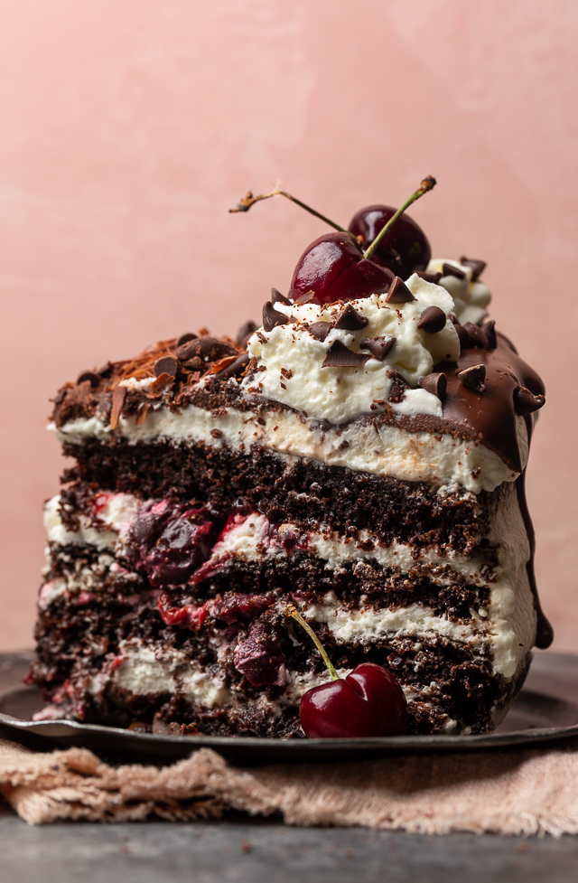 A slice of black forest cake on a pewter plate with a pink napkin and fresh cherry on the side.