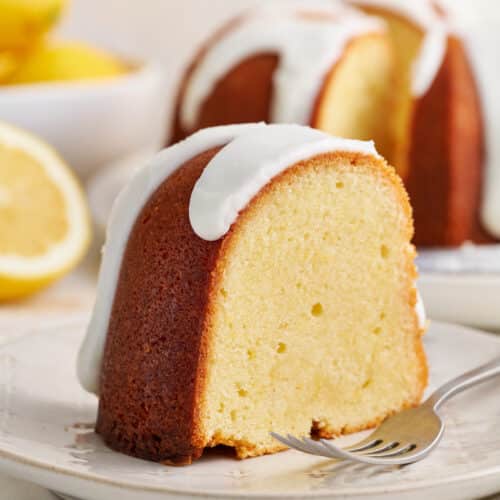 Slice of lemon bundt cake on white plate with fork and lemon slices.