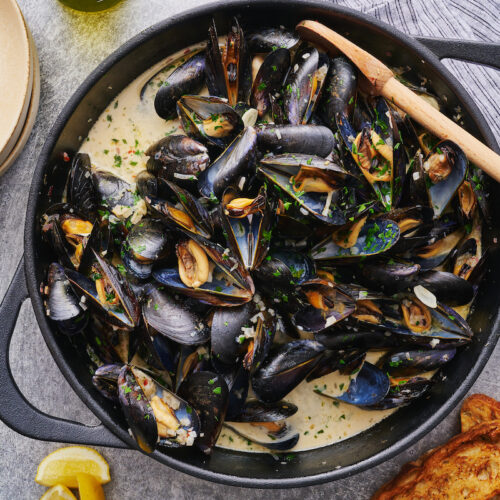 Pot of cooked fresh mussels in white wine sauce with lemon juice and white wine on the side.