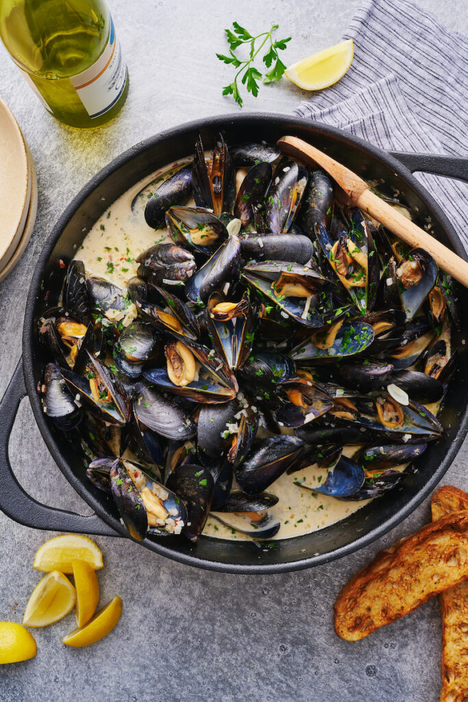 Pot of cooked fresh mussels in white wine sauce with lemon juice and white wine on the side.