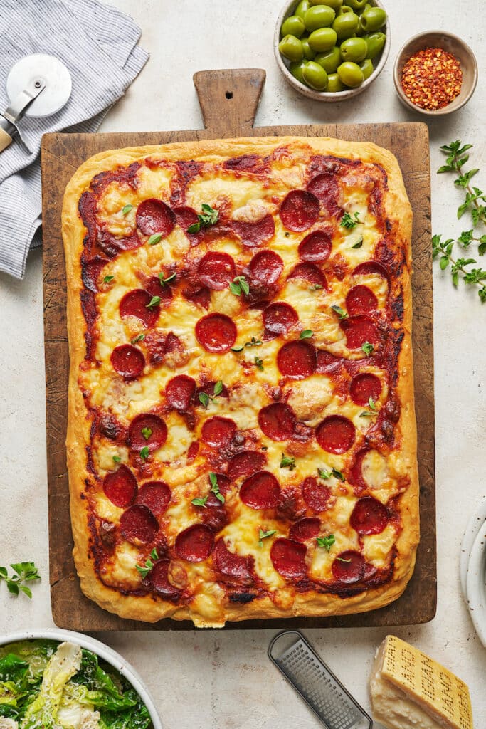 Baked pepperoni pizza on wooden cutting board with olives and salad on the side.
