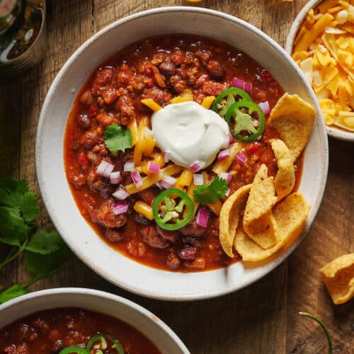 Bowl of chili topped with green onion, sour cream, and corn chips.