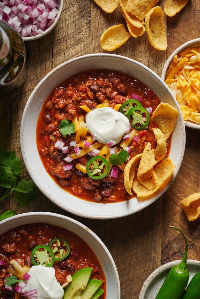 Bowl of homemade chili recipe topped with green onion, sour cream, and corn chips.
