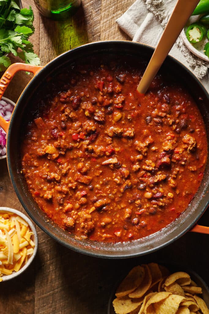 A dutch oven with homemade chili in it. Chili toppings for the chili recipe are on the brown table below.