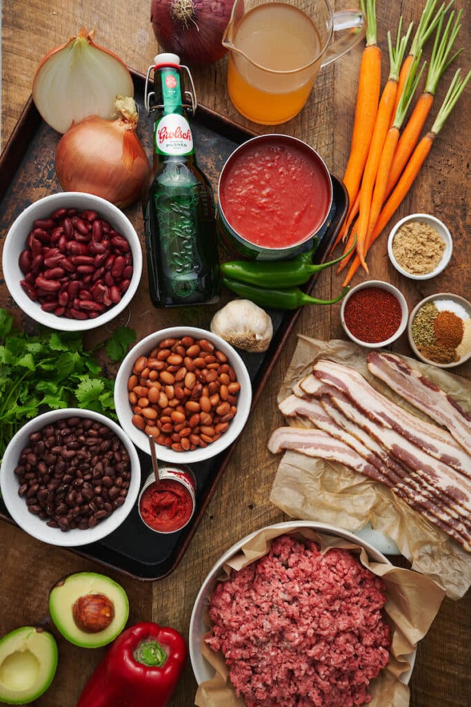 Ingredients for homemade chili recipe on a table, including black beans, kidney beans, pinto beans, ground beef, and diced tomatoes.