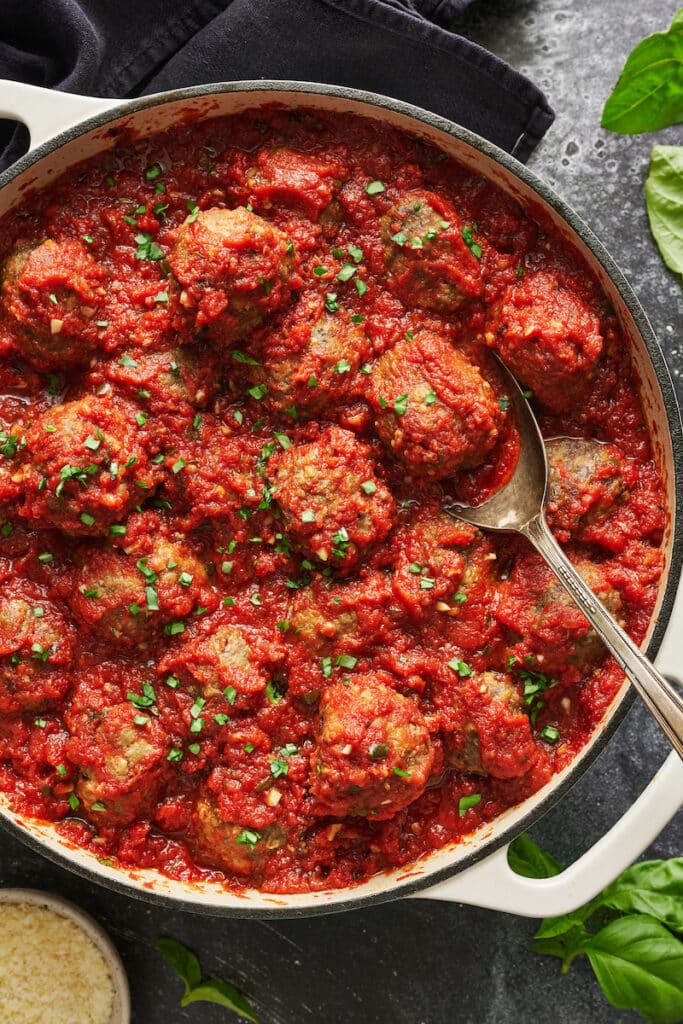 Meatballs in marinara sauce in a white pot with basil on top.