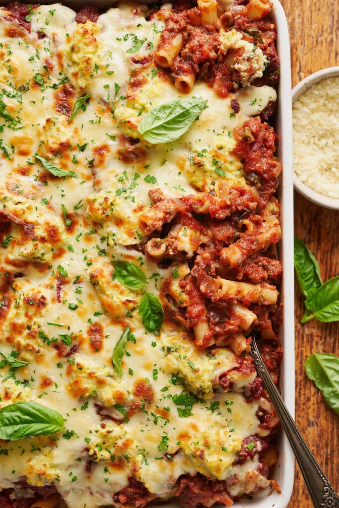 Baked ziti in casserole dish with cheese and basil on the side.